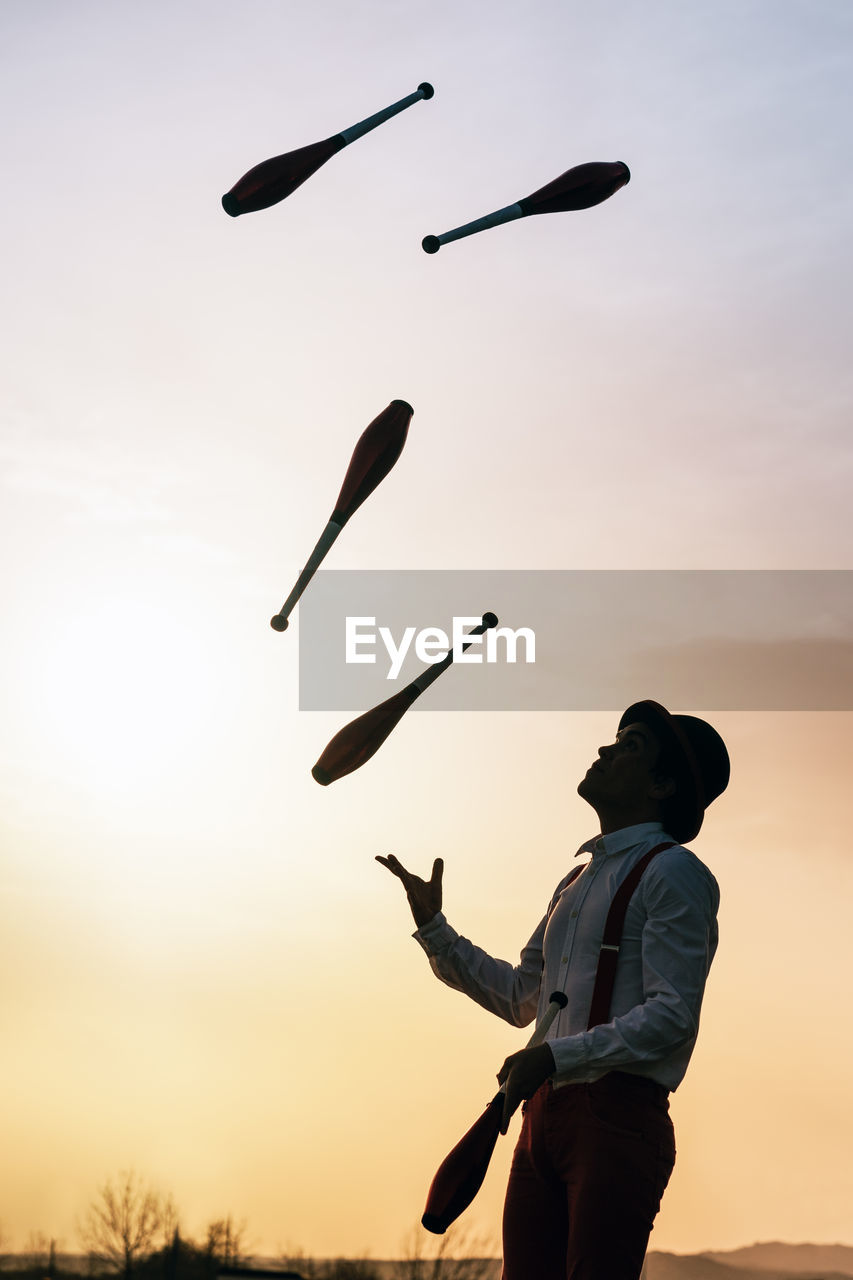 Low angle side view of unrecognizable male circus artist in hat juggling clubs against sunset sky