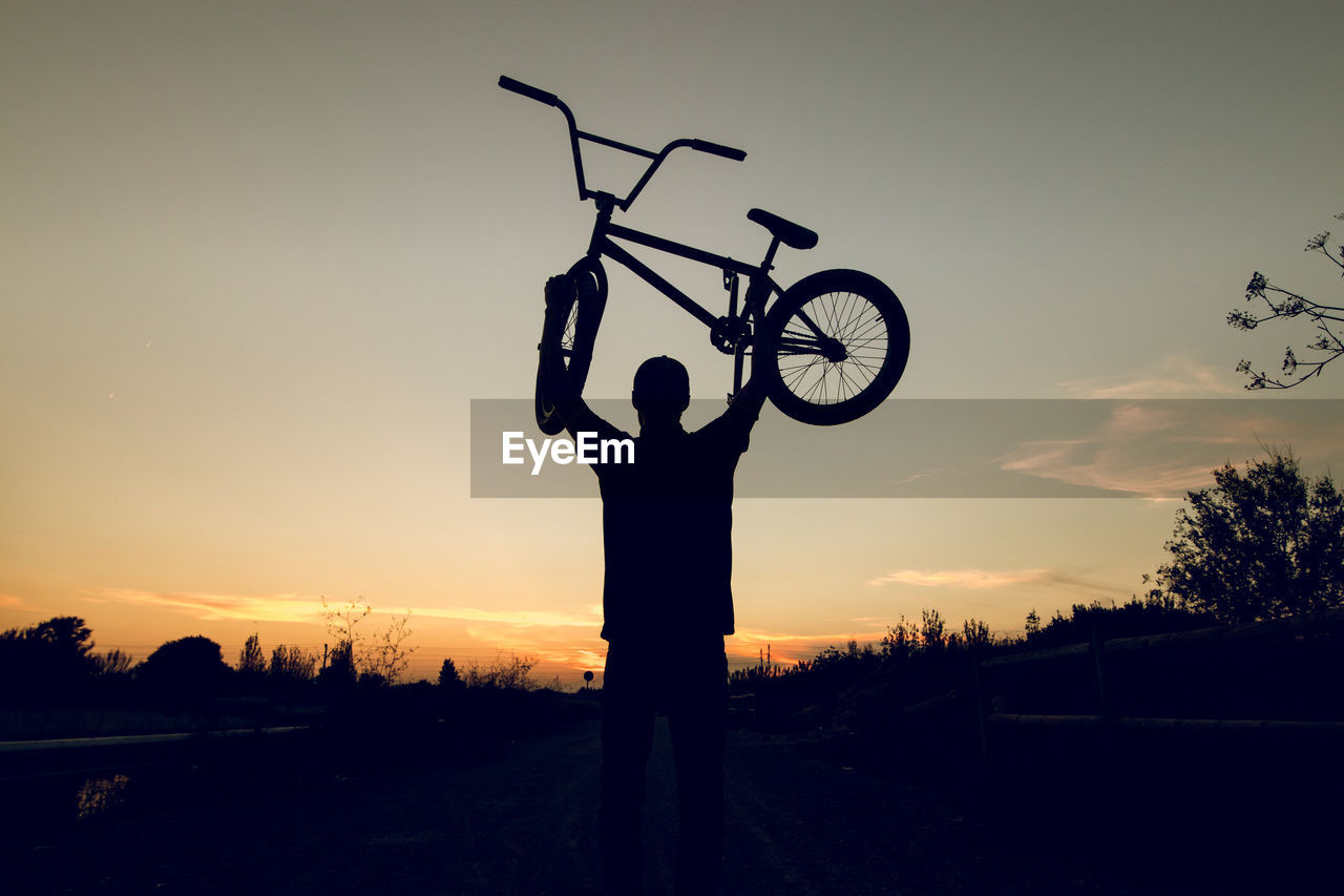 Rear view of man lifting bicycle against sky during sunset