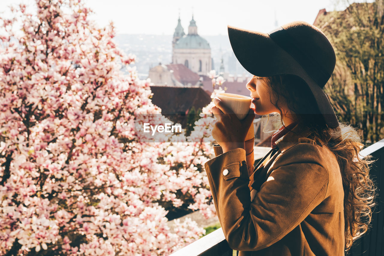 Beautiful woman in brown hat drinking coffee at outdoor cafe with amazing view of spring prague.