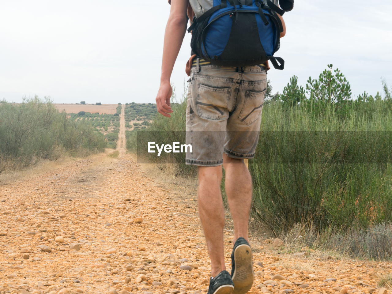Low section of man walking on dirt road against sky