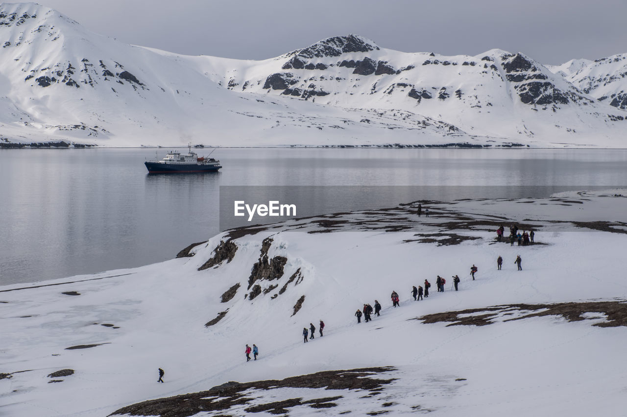 Group of explorers walking on snowcapped shoreline of arctic ocean with ship in the background.
