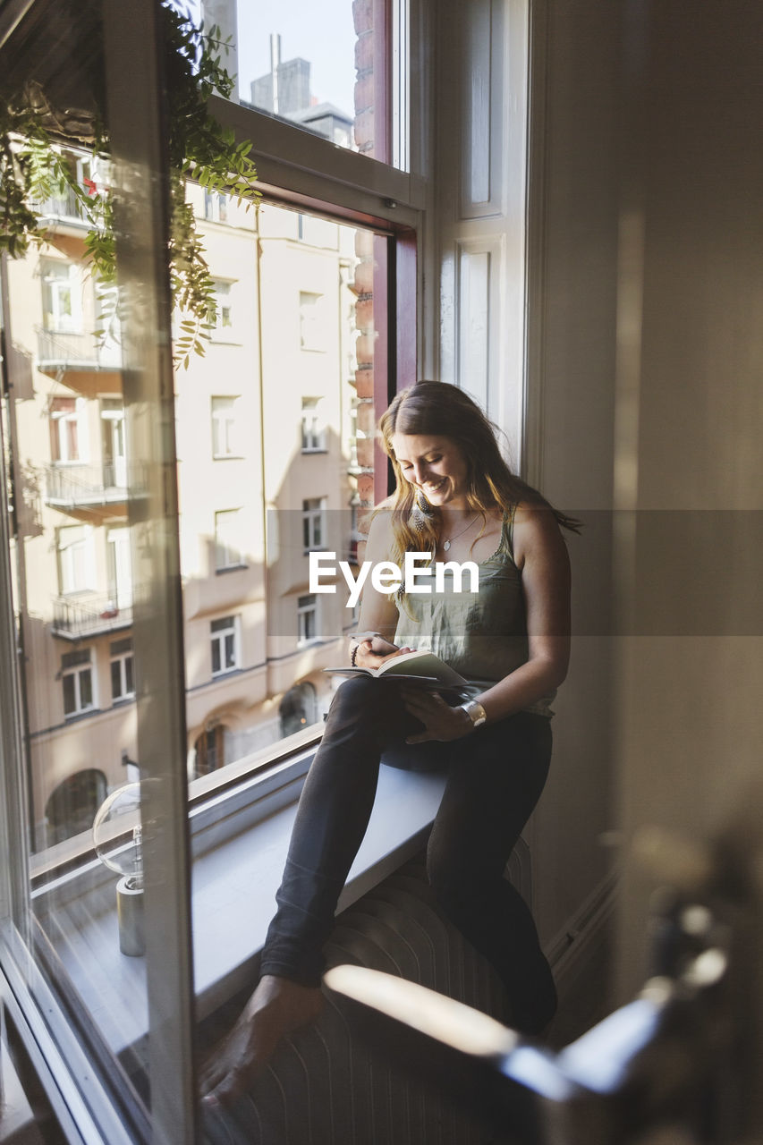 Happy woman using smart phone while holding guidebook on window sill at home