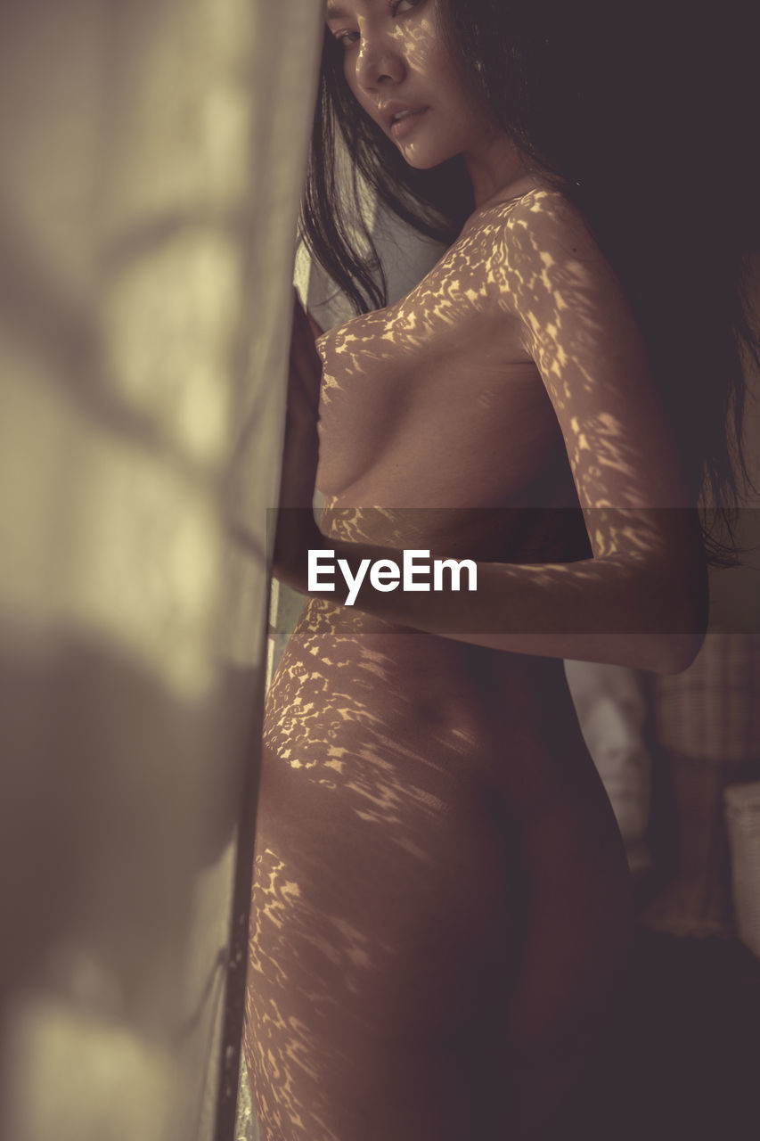 Portrait of naked young woman standing by window at home