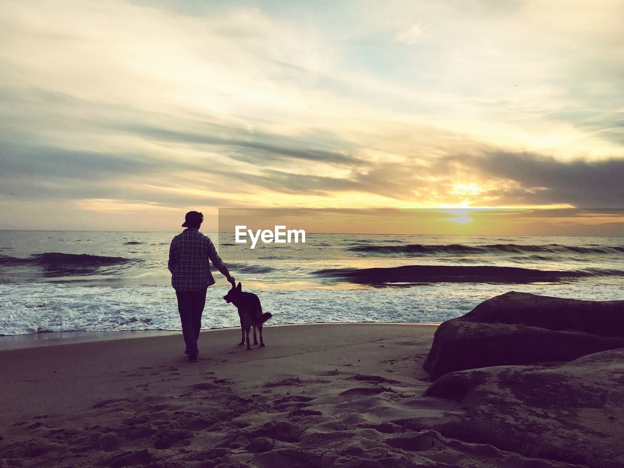 Rear view of man with dog at beach against sky during sunset
