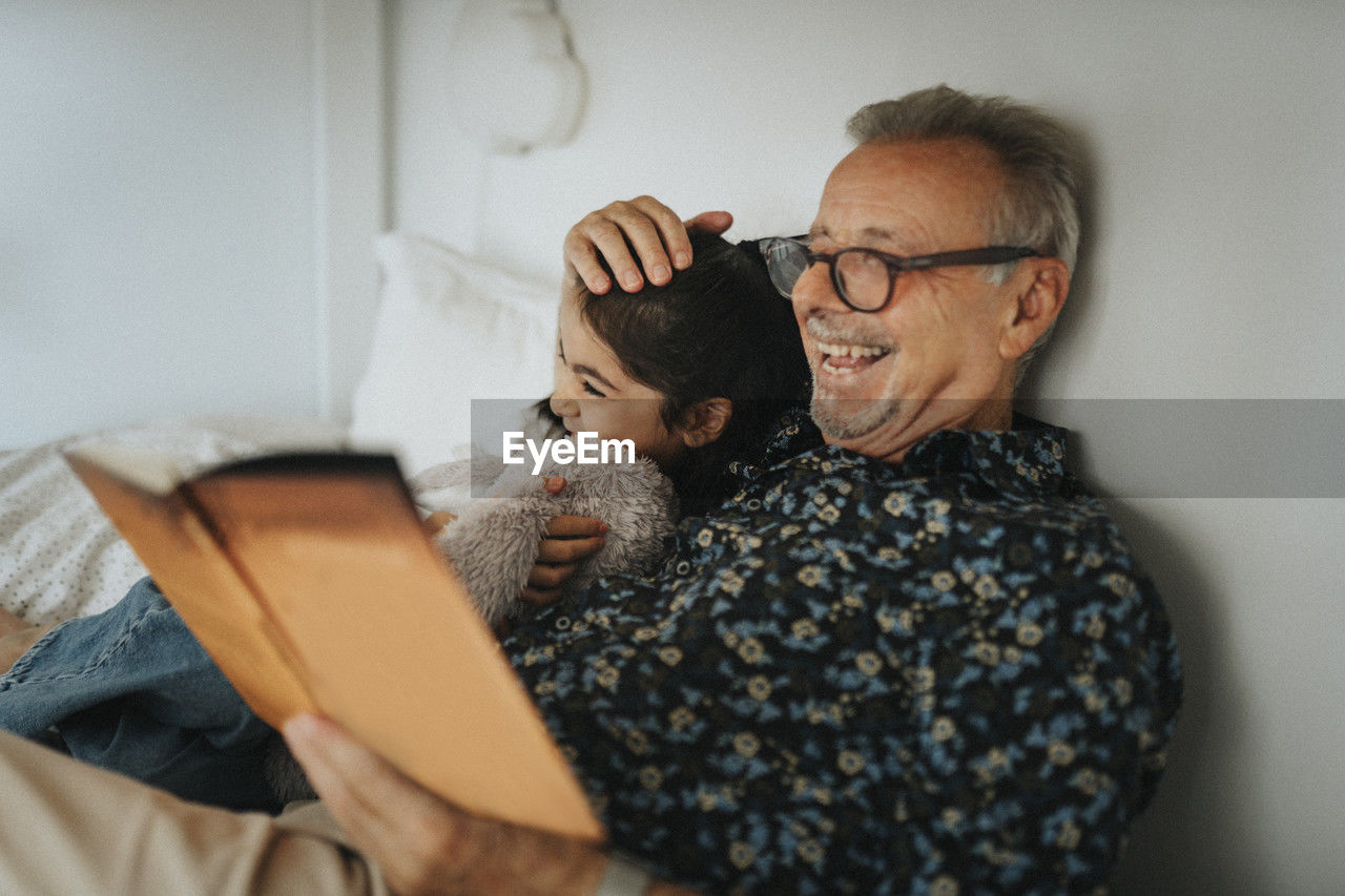 Cheerful senior man reading storybook with granddaughter on bed at home