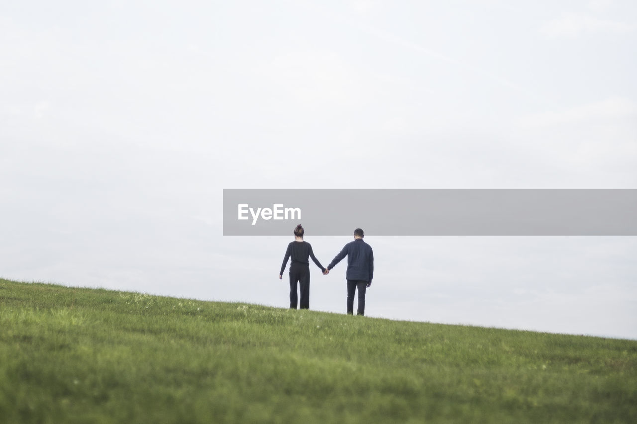 Rear view of couple holding hands standing on grassy land against sky