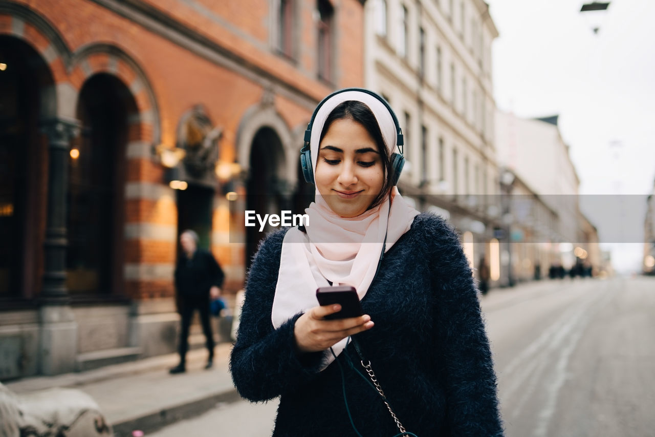 Smiling teenage girl listening to headphones using smart phone while walking on street in city