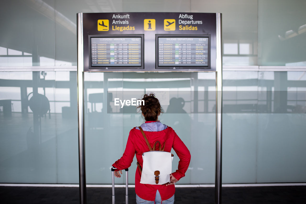 Back view of anonymous female tourist with backpack and suitcase standing near signboard with flight schedule in airport