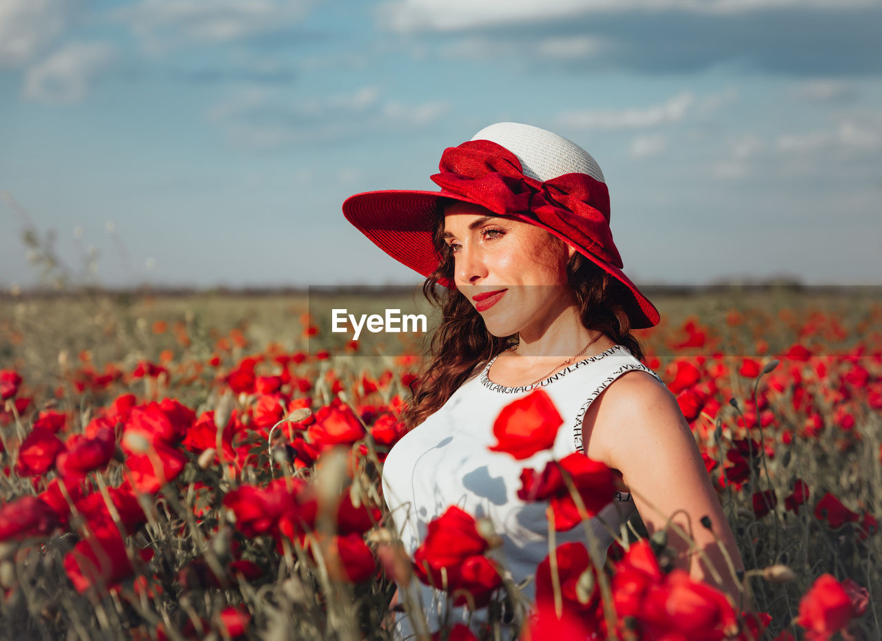 PORTRAIT OF SMILING YOUNG WOMAN STANDING BY RED POPPY FLOWERS