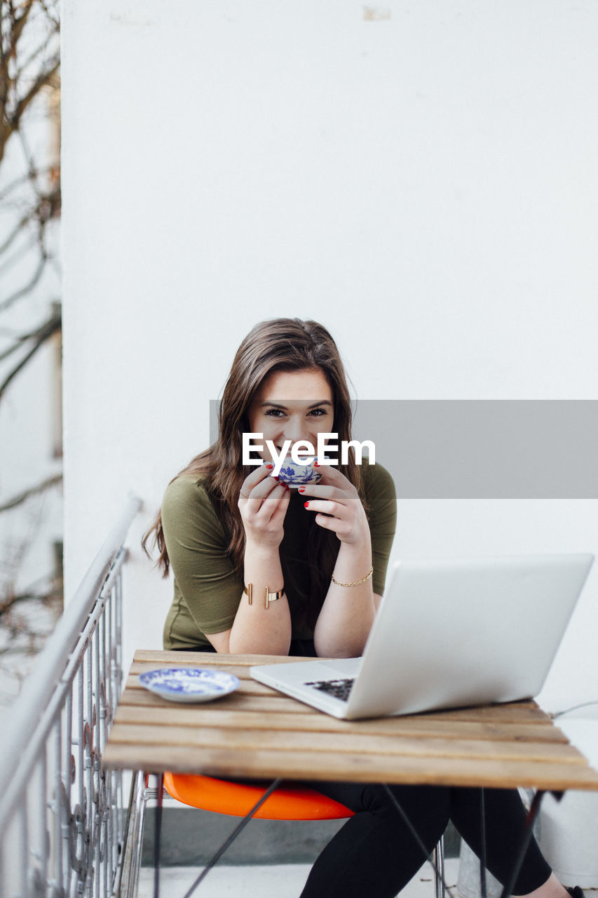 Portrait of smiling young woman with laptop on table having coffee at balcony