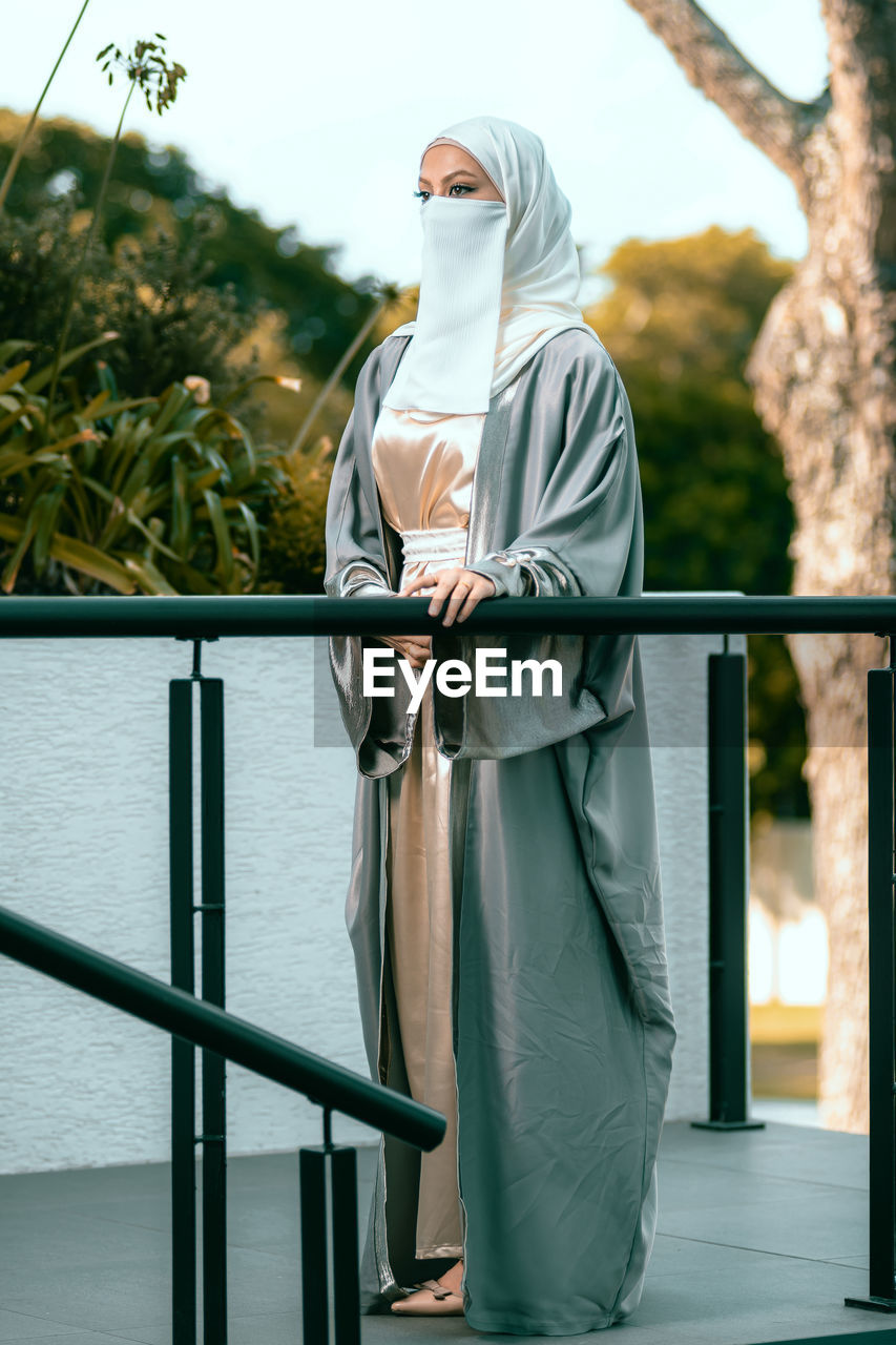 Full length picture of a veiled woman with white niqab and long dress standing against railing