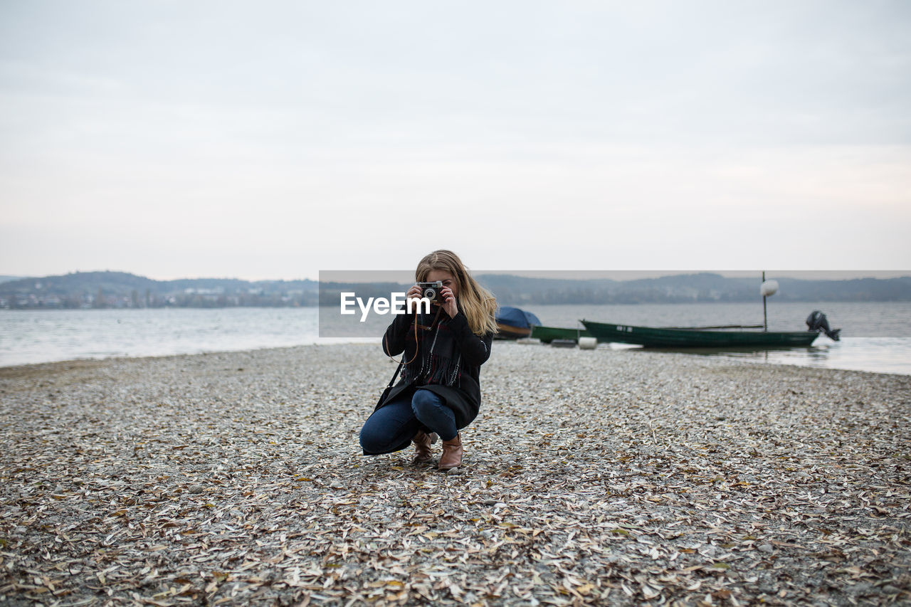 Woman photographing through camera while crouching at lakeshore against clear sky