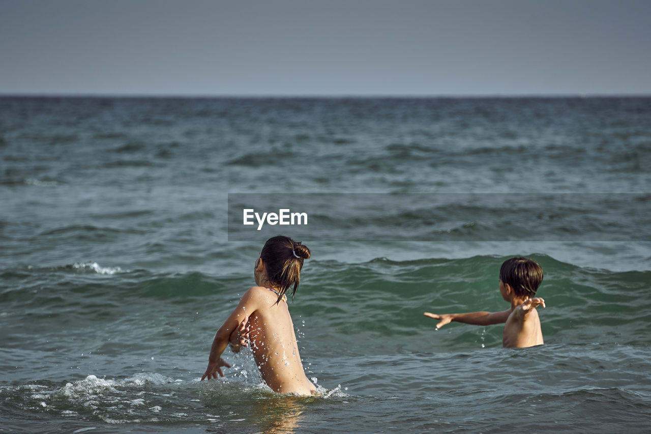 Shirtless siblings enjoying in sea against sky