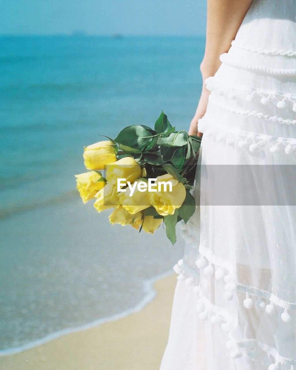 Midsection of woman holding yellow roses while standing at beach