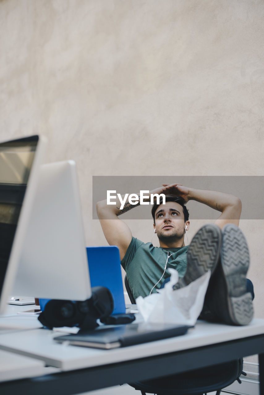 Thoughtful male computer programmer sitting with hands behind head and feet up at desk against beige wall in office