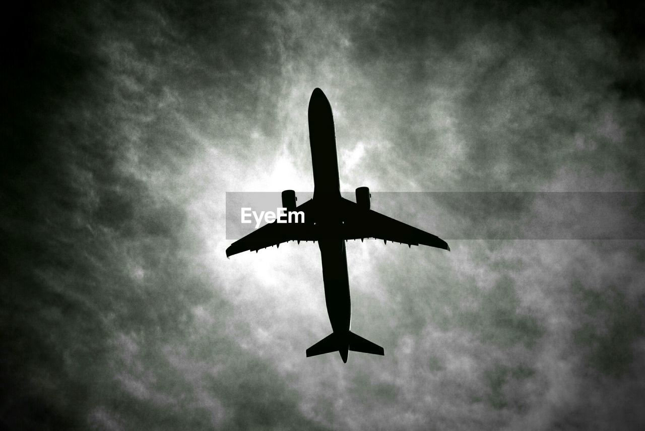 Directly below shot of silhouette airplane flying against cloudy sky