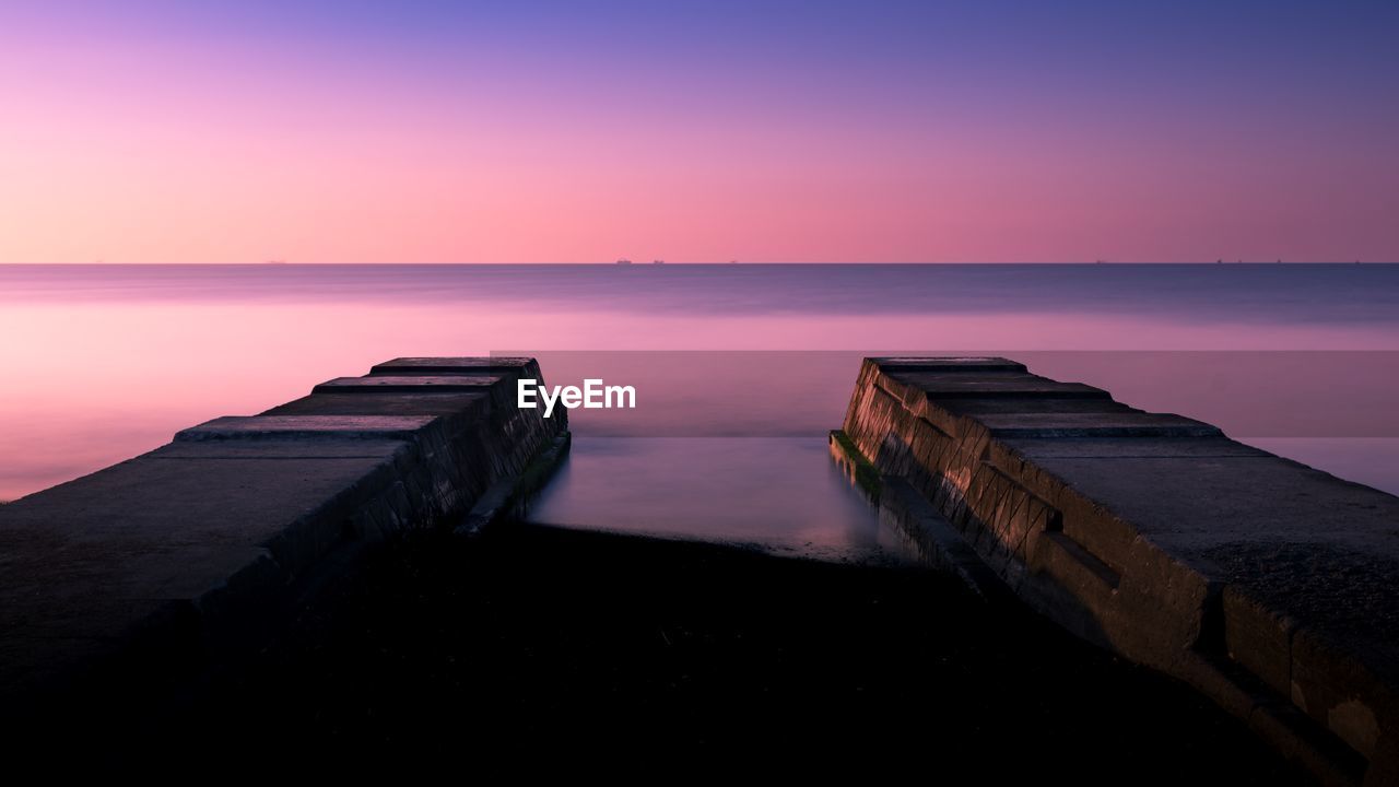 WOODEN POSTS ON SEA AGAINST SKY DURING SUNSET