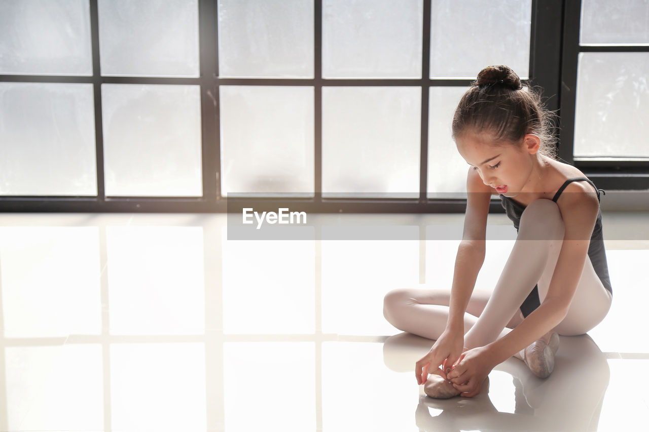Ballet tying shoe lace while sitting on floor by window