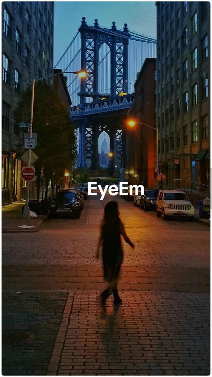 Woman walking amidst building against manhattan bridge at dusk