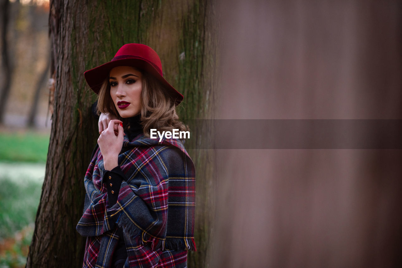 Portrait of woman wearing hat standing against tree trunk