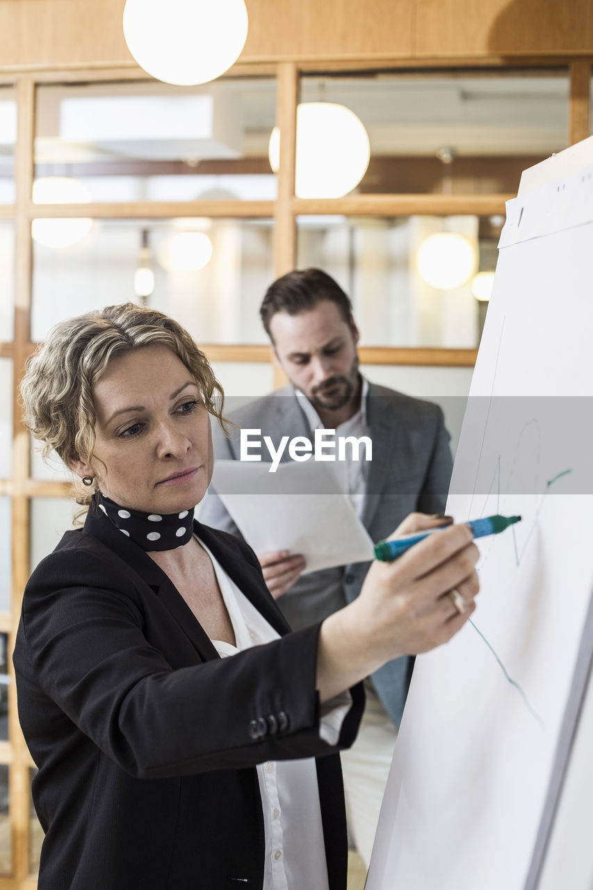 Mature businesswoman writing on flip chart with male colleague in background at office
