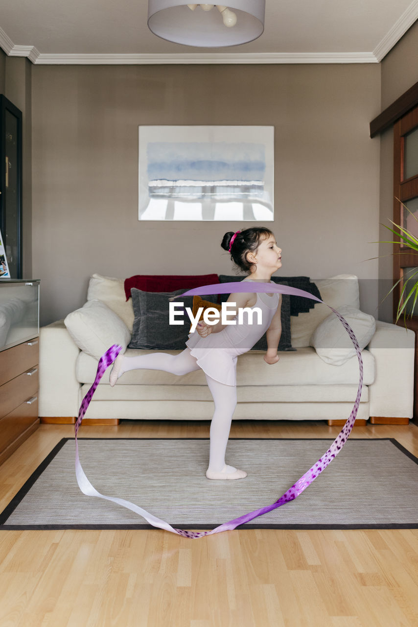 Side view of cute little girl in leotard and tights spinning ribbon and dancing during rhythmic gymnastic training in cozy living room at home