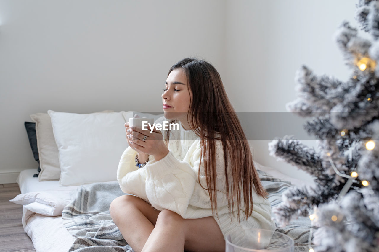 Woman in warm white winter sweater lying in bed at home at christmas eve holding cup