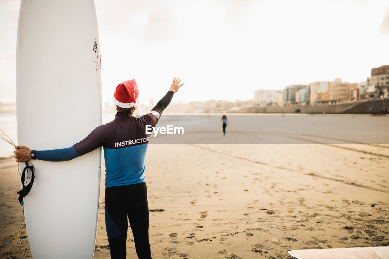 Rear view of man wearing santa hat holding surfboard at beach