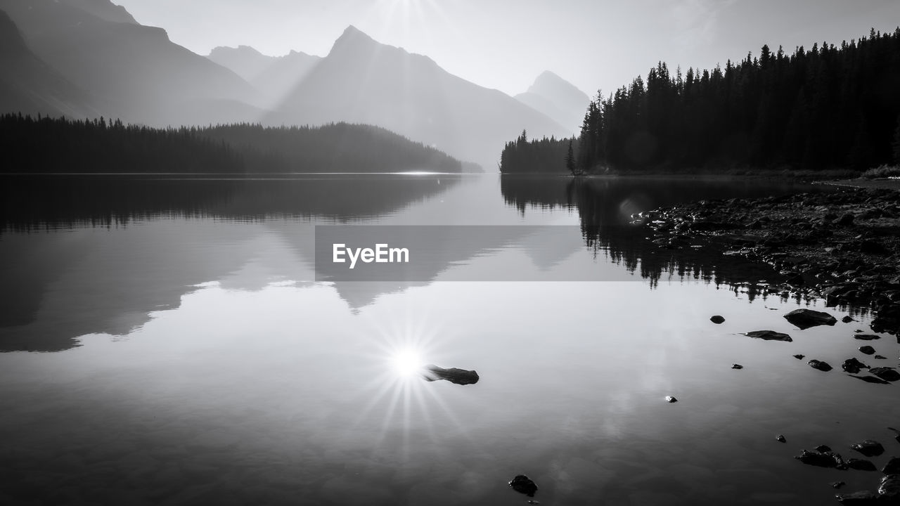 Sun reflecting in still alpine lake surrounded by mountains, monochromatic, jasper np, canada