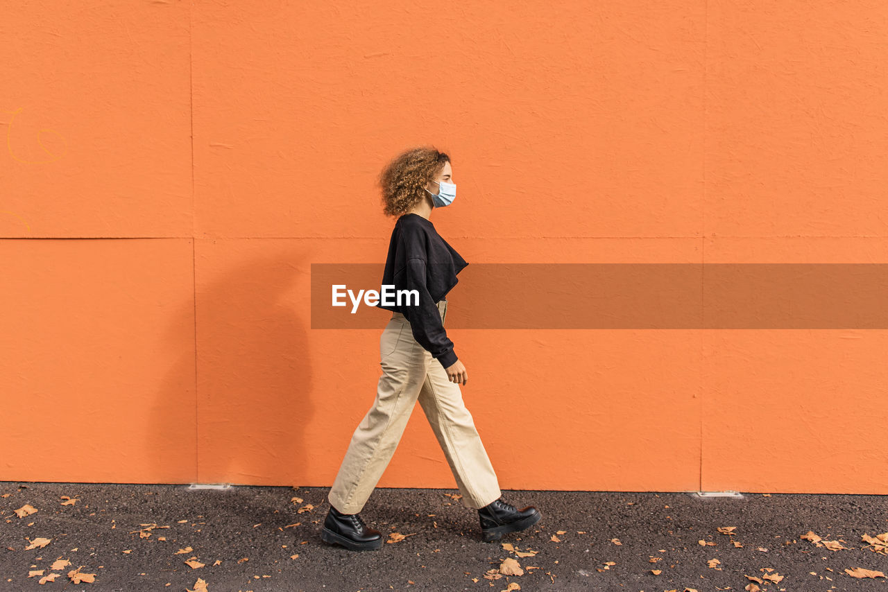 Young woman wearing face mask walking against orange wall