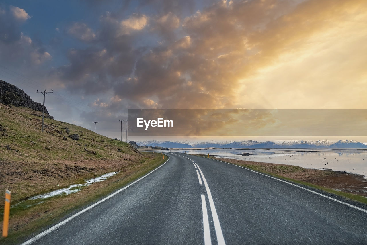 Diminishing highway amidst sea and mountain against cloudy sky during sunset