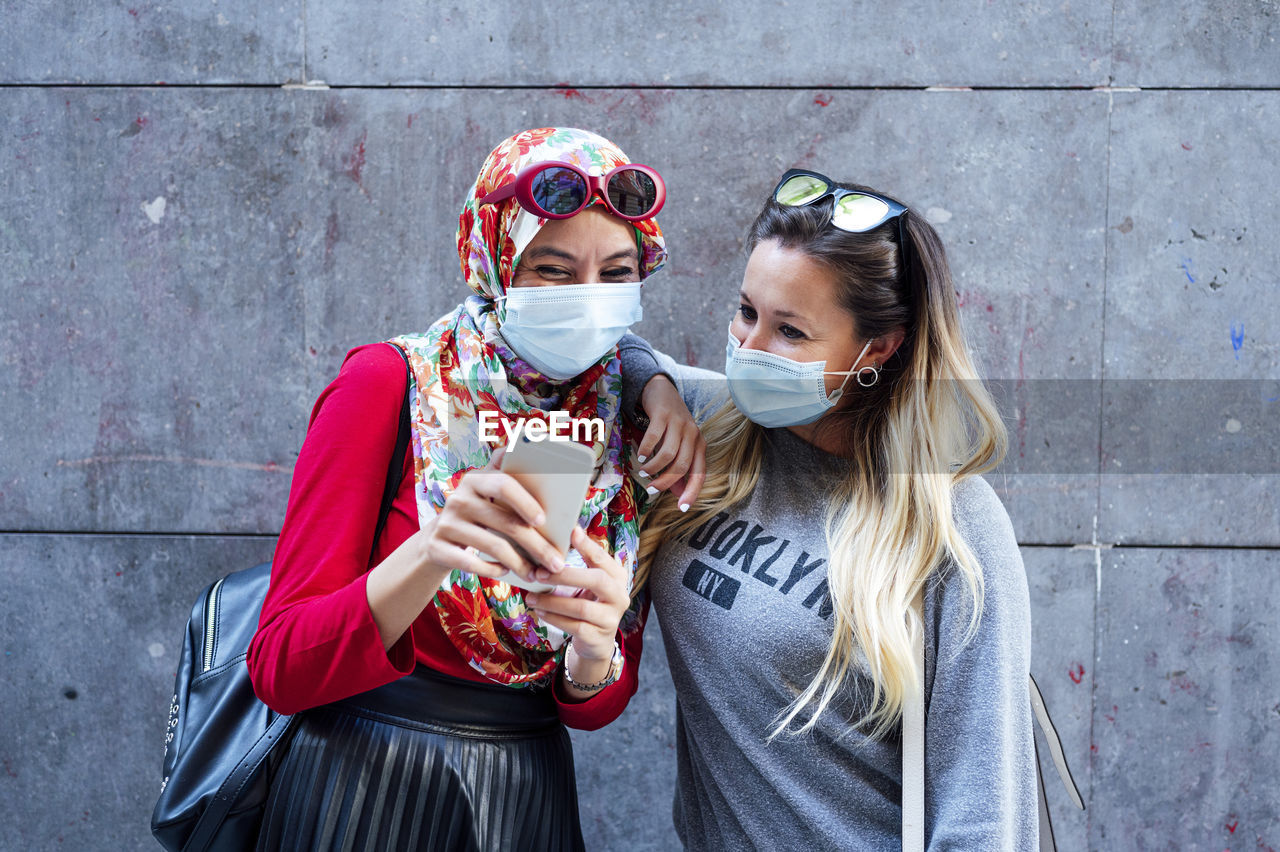 Muslim woman sharing smart phone with female friend against gray wall