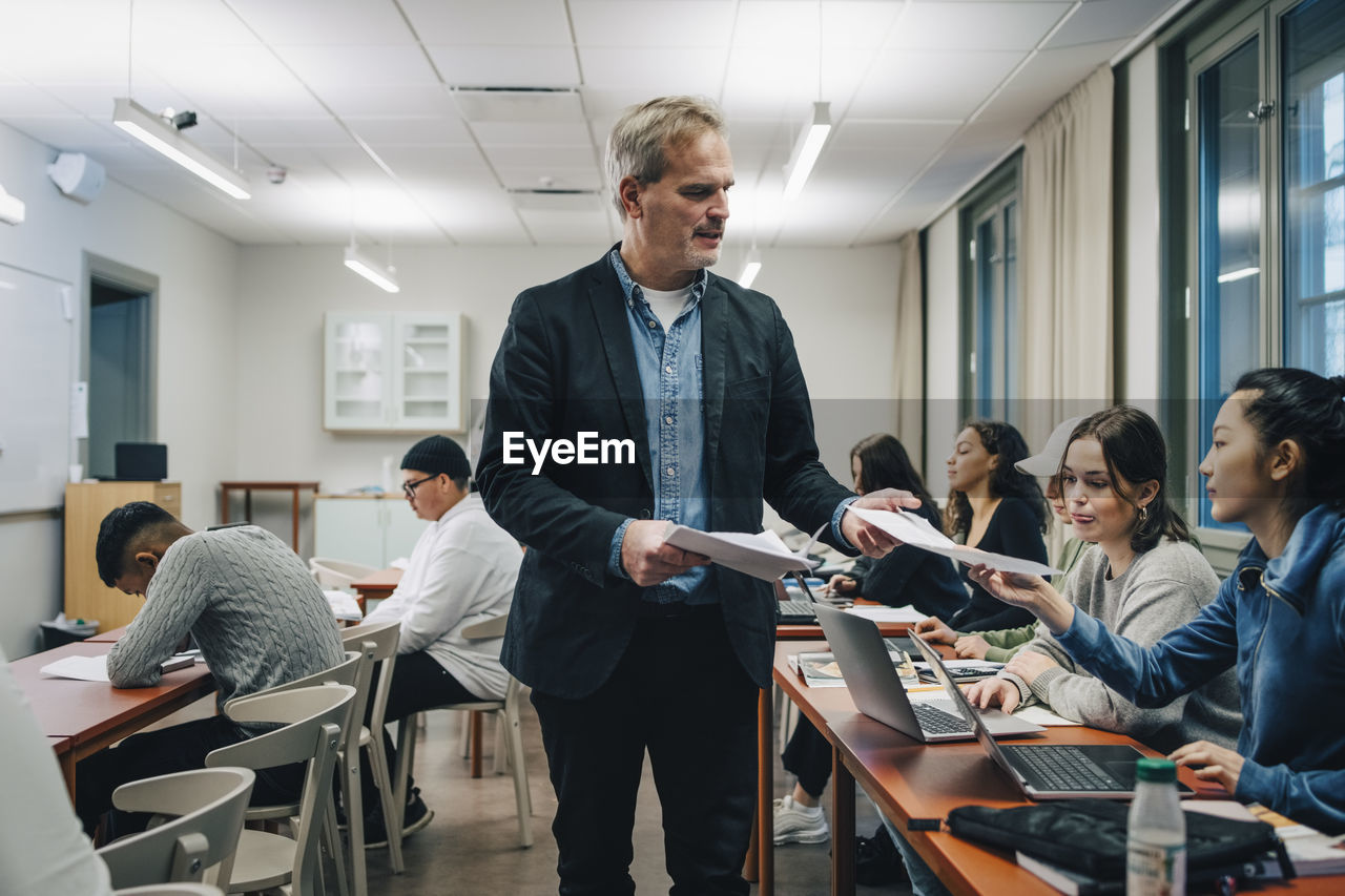 Teacher giving questionnaire to students sitting at desk in classroom