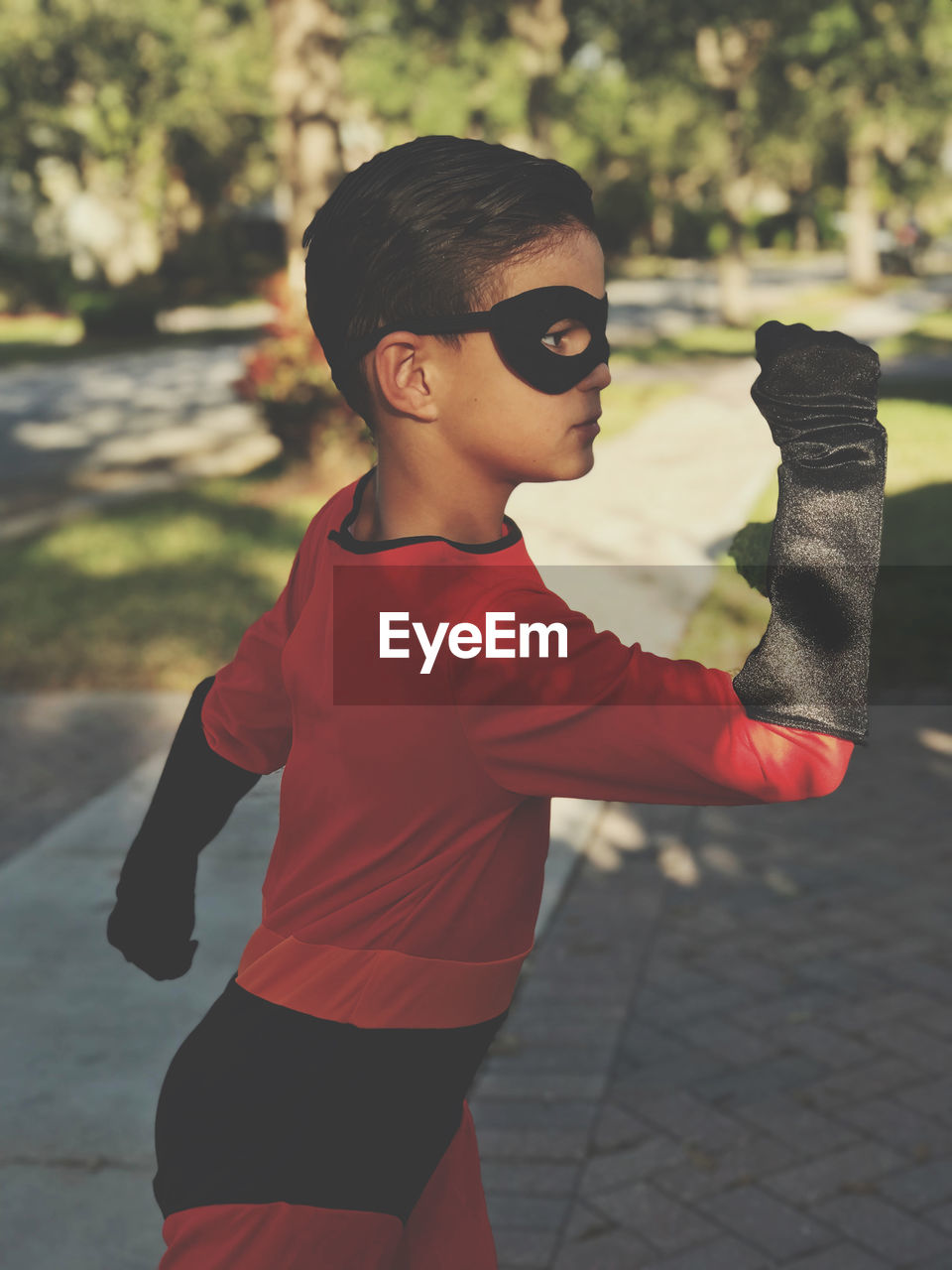 Side view of cute boy in superhero costume standing on footpath in park