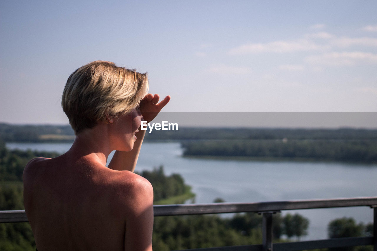 Rear view of young woman shielding eyes by railing against river