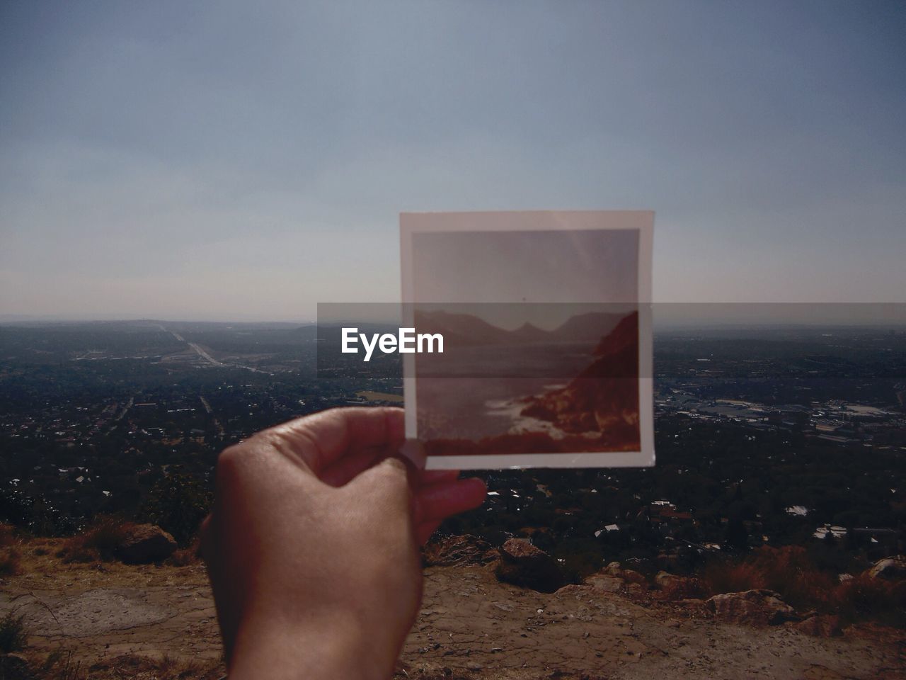 Cropped image of person holding photograph by landscape against sky