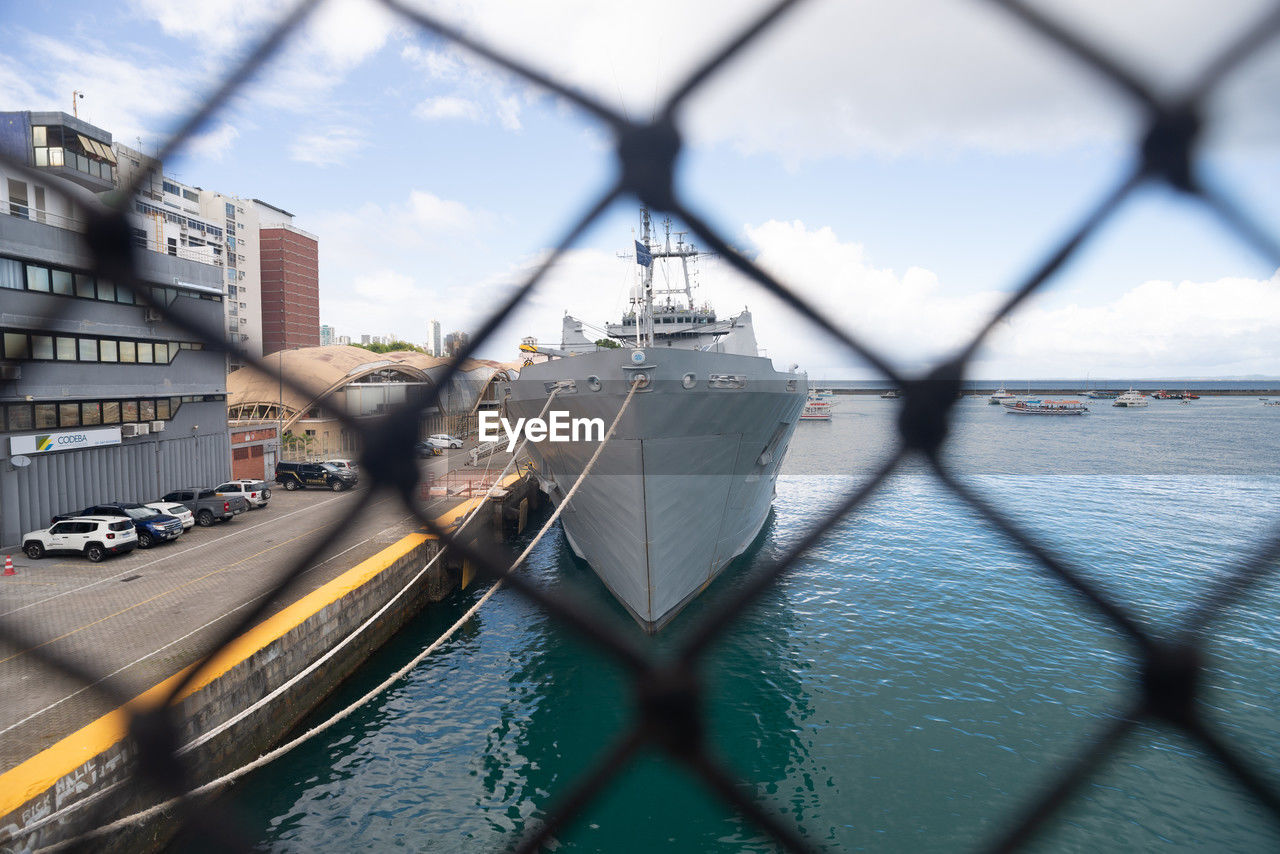 chainlink fence, fence, architecture, transportation, vehicle, built structure, mode of transportation, water, sky, city, building exterior, nature, cloud, day, security, no people, protection, nautical vessel, reflection, ship, travel destinations, outdoors, travel, building, bridge, wire mesh, blue, motion, wire, metal, focus on background, boat