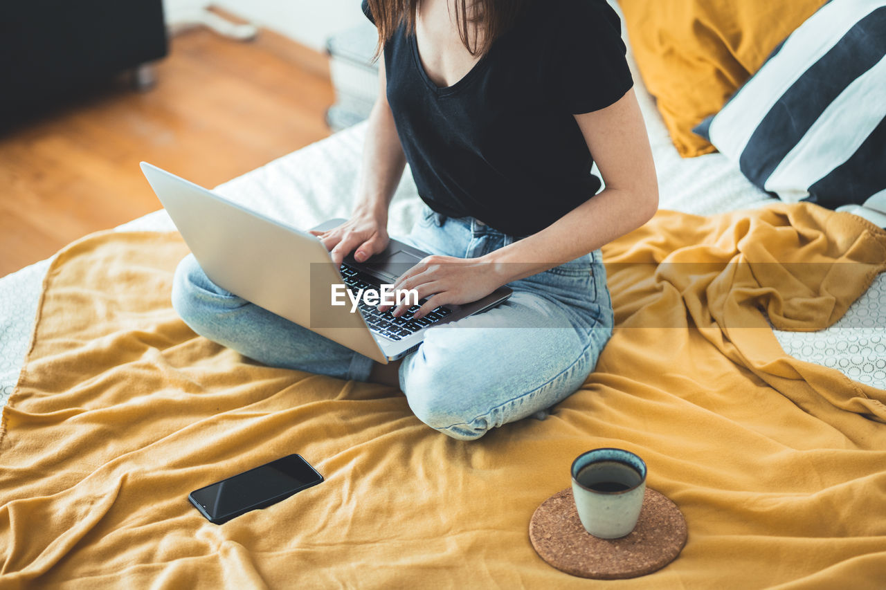 High angle view of woman using laptop on bed at home  female relaxing and drinking cup of hot coffee