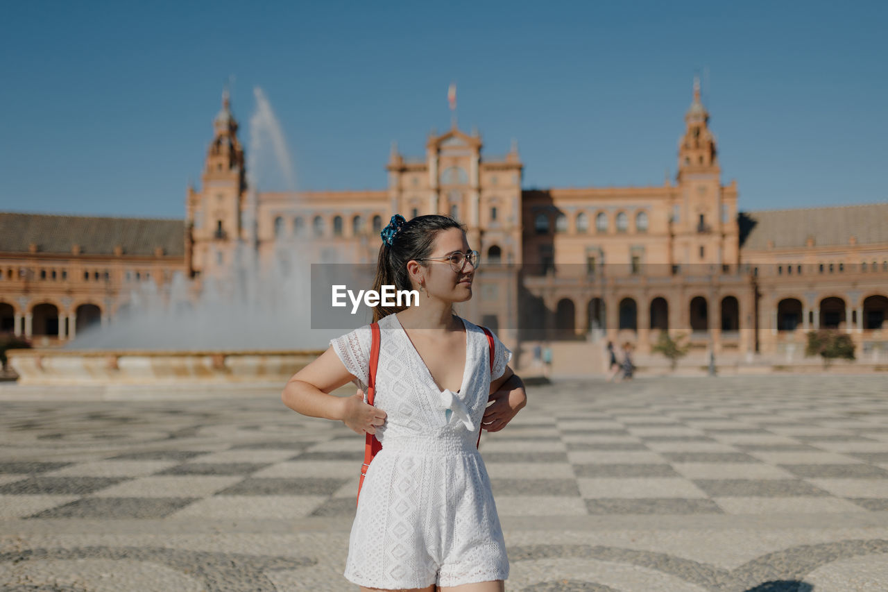 Young tourist woman wearing a white dress is looking away and smiling while standing in seville.