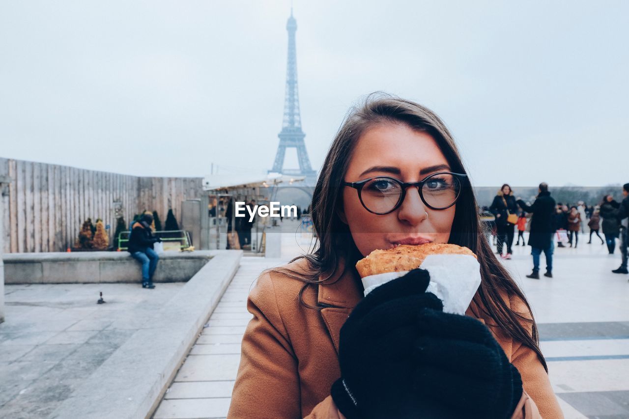 Portrait of young woman eating while standing against eiffel tower in city