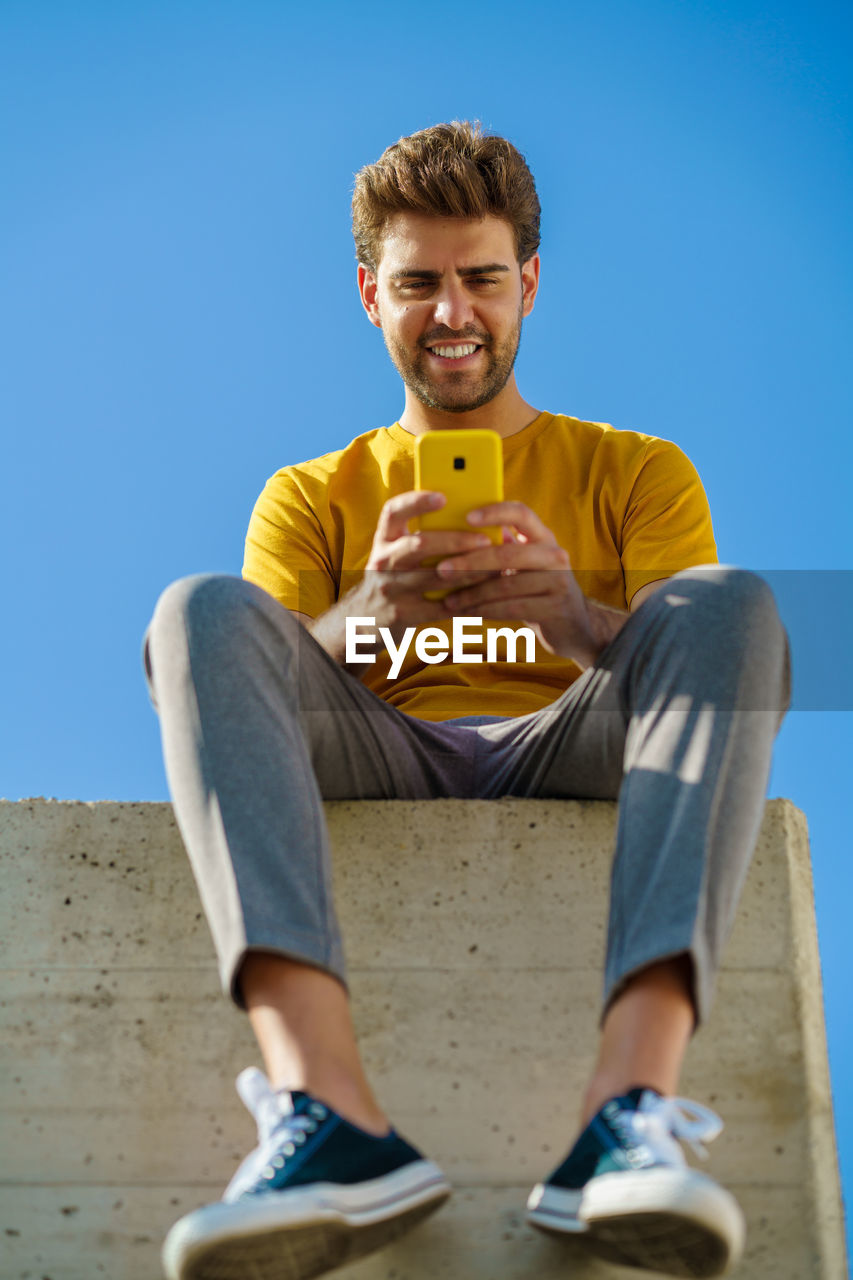 Low angel view of smiling young man using smart phone sitting against sky