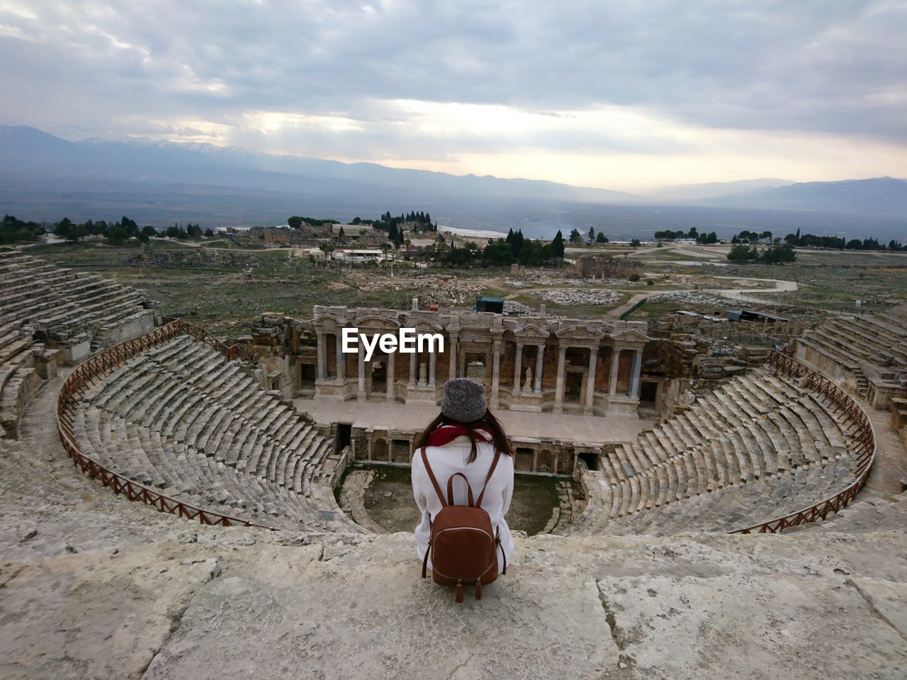 Rear view of woman sitting at amphitheater against cloudy sky during sunset