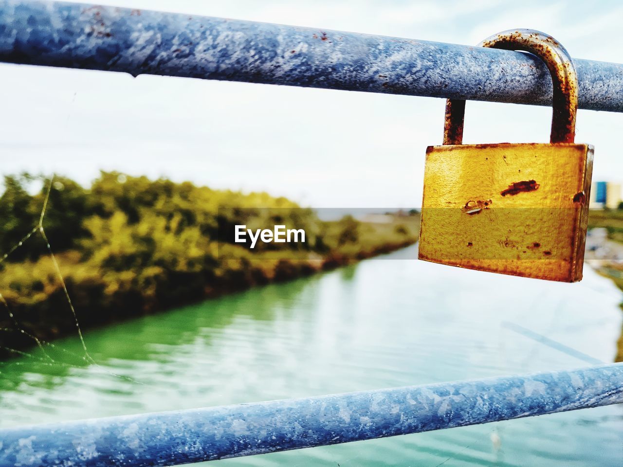 water, padlock, yellow, protection, blue, security, lock, nature, metal, hanging, focus on foreground, railing, no people, day, river, outdoors, love, positive emotion, close-up, green, hope, emotion, fence, reflection, tree, sky, bridge