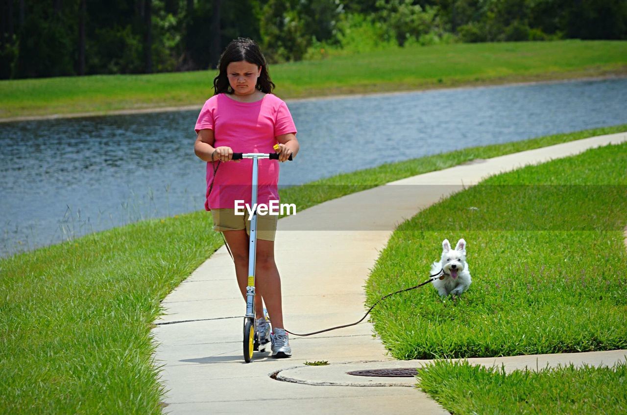 Girl riding on push scooter by dog running on grassy field against lake at park