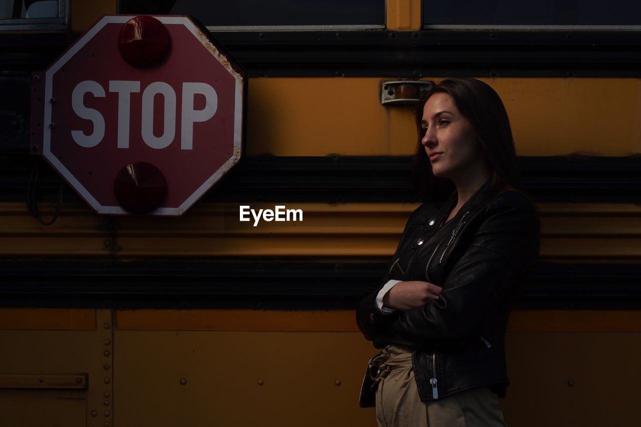 Young woman looking away while standing against bus at night