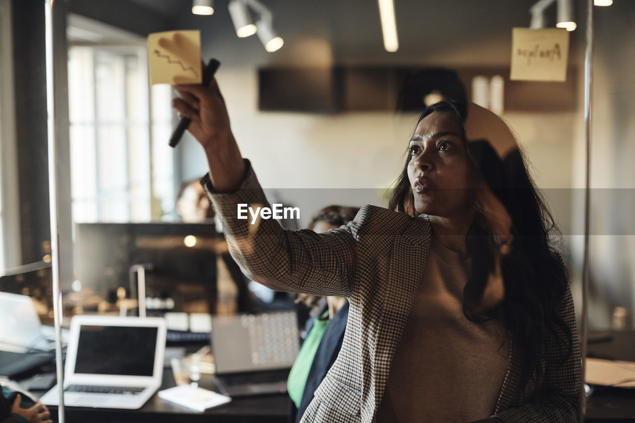 Mature businesswoman sticking adhesive note on glass wall in office