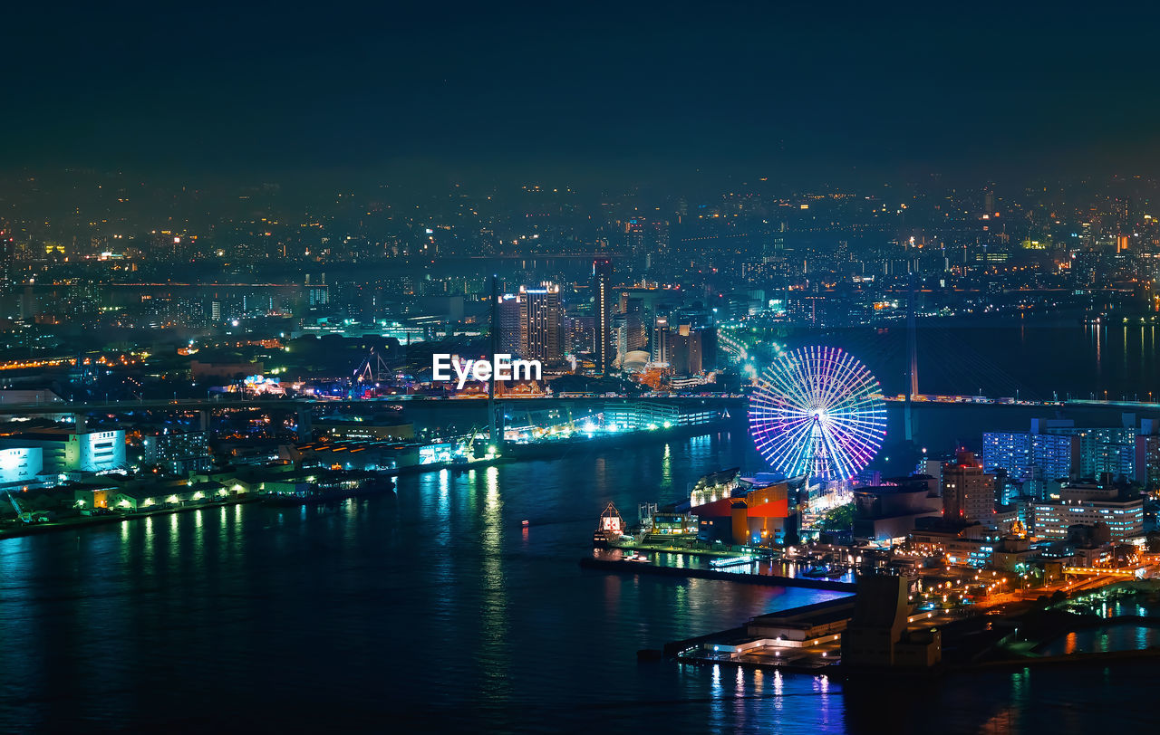 Illuminated ferris wheel by river in city against sky at night