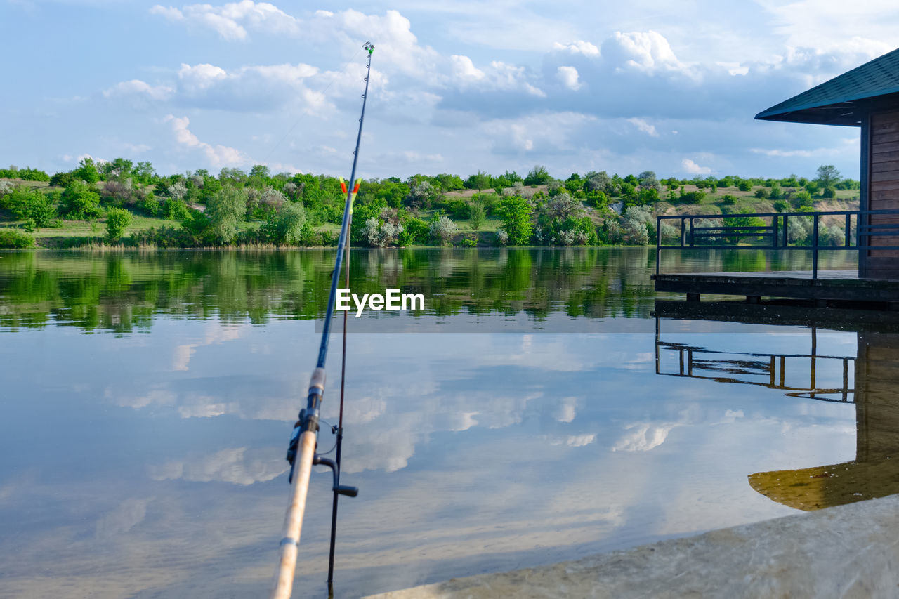 Fishing rod on a pier by the river. spring sunny day, beach with trees, sky reflection in the water.