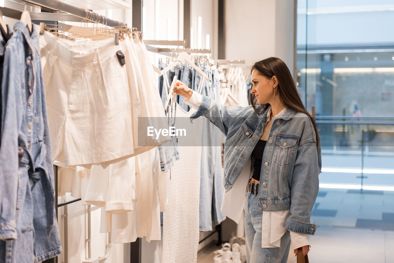 Young woman stands in a fashion store, carefully choosing clothing items to buy. she browses through