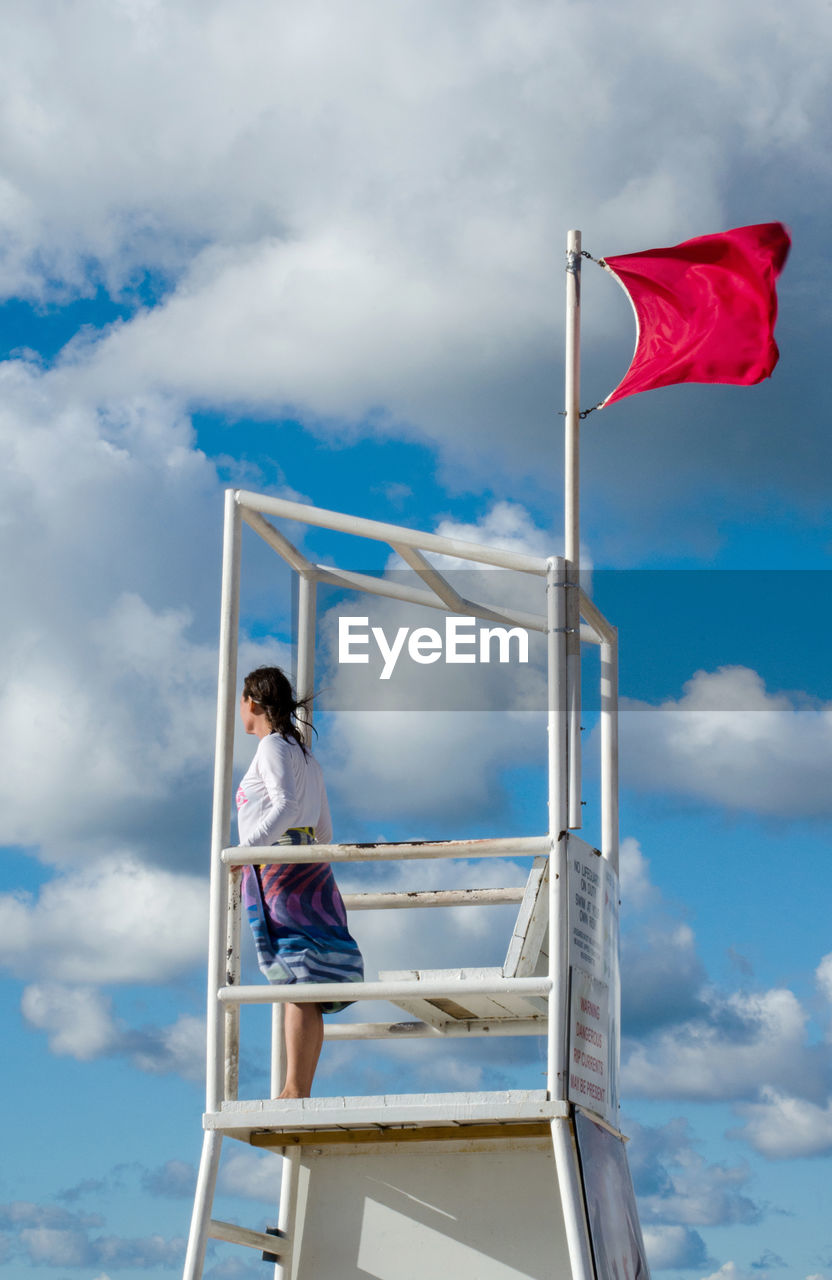 Young woman watches the water from a life guard tower on the beach, while a red flag flies 
